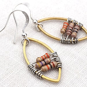 May include: Unique handmade earrings featuring a gold-toned oval frame with silver wire accents. Each earring showcases three colorful resistor beads, creating a distinctive and artistic design. The earrings have silver ear wires.