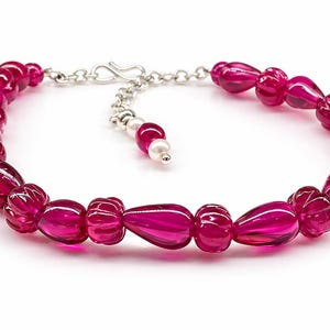 Könnte beinhalten: Ein magentafarbenes Armband mit Silberverschluss und Kette. Das Armband hat durchscheinende, tropfenförmige Perlen. Eine kleine Kette mit einer Perle und einer magentafarbenen Perle hängt am Verschluss.