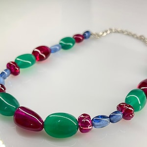 Puede incluir: Un collar de cuentas de colores con cuentas ovaladas y redondas verdes, magenta y azules. El collar tiene un cierre de cadena plateada. Una joya vibrante y llamativa.