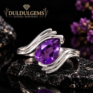 Op de afbeelding: Een zilveren ring met een peervormige paarse amethist, gezet tussen twee golvende, gepolijste zilveren banden. De tekst DULDULGEMS NATURAL GEMSTONES is bovenaan zichtbaar. De ring heeft een gladde, metalen afwerking en een levendige paarse kleur.