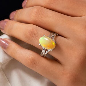 May include: A silver ring featuring a large, oval-shaped cabochon opal with iridescent yellow and orange flashes. The band has a split-shank design that tapers toward the center setting, which holds the gemstone securely with four small prongs.