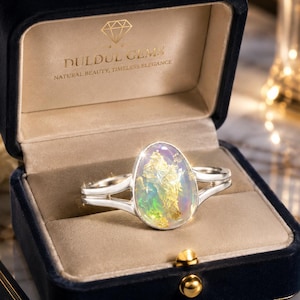 Könnte beinhalten: Ein Silberring mit einem großen, ovalen, schillernden Opalstein mit Goldeinschlüssen, gefasst in eine polierte Silberschiene. Der Ring befindet sich in einer dunkelblauen Samtschmuckschatulle mit dem in Gold gedruckten Text DULDUL GEMS.