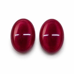 May include: Two oval-shaped, polished gemstones with a deep red hue. The gems have a smooth, reflective surface, with a horizontal white highlight across the center of each stone. The background is plain white.