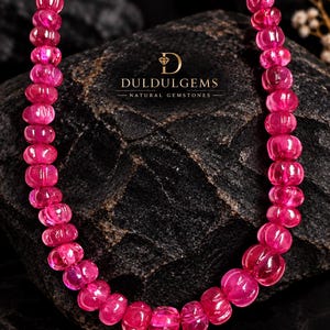 Puede incluir: Un collar que presenta una hilera de cuentas de piedras preciosas translúcidas, de color rosa vibrante, redondas y acanaladas. Las cuentas están ensartadas en un bucle continuo. El texto DULDULGEMS NATURAL GEMSTONES con un icono de diamante es visible.