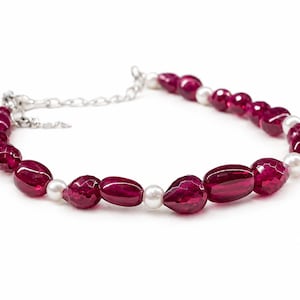 Op de afbeelding: Een armband met gefacetteerde, dieprode edelstenen en witte parels. De armband is geregen op een zilveren ketting en sluiting. De edelstenen hebben een gepolijst, reflecterend oppervlak. De parels zijn rond en glad. Een sieraad.