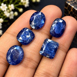 Könnte beinhalten: Fünf facettierte blaue Saphire in verschiedenen Formen, darunter ovale, kissenförmige und rechteckige Schliffe. Die Edelsteine weisen tiefe Blautöne mit internen Einschlüssen und reflektierenden Oberflächen auf und sind auf einer flachen Oberfläche angeordnet.