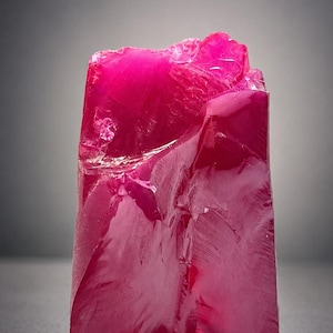 May include: A vibrant, rectangular-shaped, raw ruby gemstone. The gem is a deep, saturated pink color with a slightly rough, textured surface. The ruby is standing upright against a dark gray background.