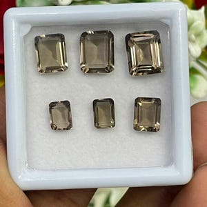 Puede incluir: Una caja de presentación rectangular blanca que contiene seis piedras preciosas de cuarzo ahumado sueltas, facetadas y con corte esmeralda. Las piedras están dispuestas en dos filas, con tres piedras más grandes arriba y tres más pequeñas abajo, todas con un tono marrón translúcido.