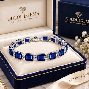 Puede incluir: Una pulsera de tono plateado con una serie de piedras preciosas rectangulares de color azul intenso engastadas en eslabones metálicos. La pulsera se presenta dentro de un joyero azul marino forrado de terciopelo con el texto DULDULGEMS NATURAL GEMSTONES.