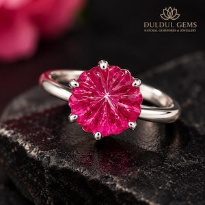 May include: A silver ring featuring a round, vibrant pink gemstone with a faceted, radial sunburst pattern. The gemstone is secured by six prongs on a smooth, polished silver band. The text DULDUL GEMS NATURAL GEMSTONES & JEWELLERY is visible.