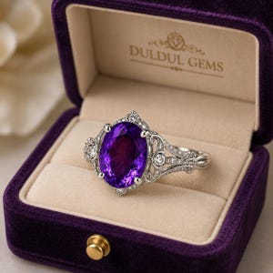 Könnte beinhalten: Ein silberner Ring mit einem großen, oval geschliffenen violetten Amethyst, umgeben von kleinen klaren Schmucksteinen. Die Ringschiene weist filigrane Metallarbeiten auf und liegt in einer violetten Samtschatulle mit dem Aufdruck DULDUL GEMS.