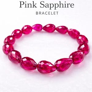 May include: A pink sapphire bracelet with teardrop-shaped gemstones. The gemstones are a vibrant fuchsia color and are strung together to form a circular shape. The text "Pink Sapphire BRACELET" is displayed above the bracelet.