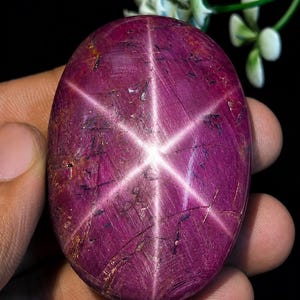 May include: An oval-shaped cabochon gemstone with a deep magenta-red colour and a prominent six-rayed star effect known as asterism. The surface displays natural inclusions and dark linear markings across the polished, translucent stone.