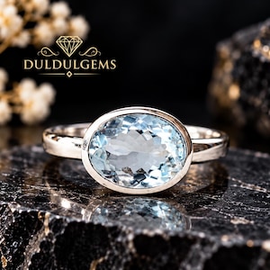 Op de afbeelding: Een zilveren ring met een grote, lichtblauwe edelsteen in ovale slijpvorm, gezet in een gladde zetting. De ring ligt op een donker, getextureerd oppervlak. De tekst DULDULGEMS is bovenaan zichtbaar, samen met een diamanten icoon.