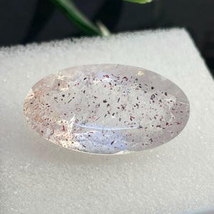 May include: An oval-shaped, faceted clear gemstone featuring internal inclusions of small, scattered red and black mineral flecks throughout the transparent stone. The polished surface reflects light, highlighting the internal texture of the crystal.