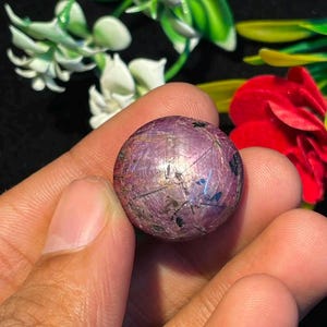 Pode incluir: Uma pedra preciosa cabochão redonda e polida com um tom magenta e roxo profundo. A superfície exibe um fenómeno ótico natural em forma de estrela conhecido como asterismo, caracterizado por linhas que se cruzam e inclusões escuras.
