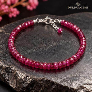 Può includere: Un bracciale composto da piccole perle di rubino rosa intenso sfaccettate disposte in cerchio. Il gioiello presenta una chiusura a moschettone color argento e una catenella di estensione regolabile che termina con una singola perla di rubino. Il testo DULDULGEMS è visibile.