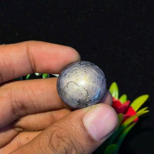 Op de afbeelding: Een ronde, in cabochon geslepen stersaffier edelsteen met een grijs-paarse tint, een zichtbaar zesstralig stereffect en donkere insluitingen over het oppervlak, met een gladde, gepolijste afwerking en een duidelijke reflecterende glans.