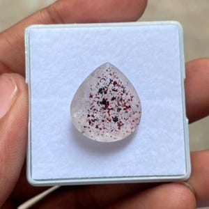 Peut inclure: Une pierre précieuse facettée en forme de poire avec une base claire et translucide, présentant de nombreuses petites inclusions dispersées dans des tons de rouge profond et de noir. La pierre est centrée sur un support en mousse blanc.