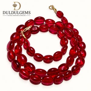 Peut inclure: Un collier composé de nombreuses perles ovales rouge foncé translucides enfilées ensemble. La pièce présente un fermoir à ressort en métal doré. Le nom de marque DULDULGEMS avec un logo en diamant est visible dans le coin supérieur gauche.