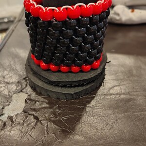 May include: A black and red beaded cuff bracelet. The bracelet is cylindrical, with black beads forming the main body and red beads outlining the top and bottom. White string is visible between the red beads. The bracelet sits on a black, layered base.