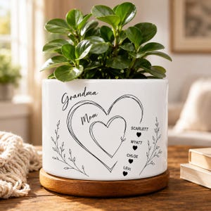 May include: A white ceramic cylindrical planter with a wooden base containing a green plant. The planter features black line art of two nested hearts, the words Grandma and Mom, and a vertical list of names Scarlett, Wyatt, Chloe, and Levi.