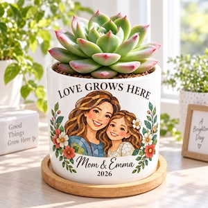 May include: A white ceramic planter on a round bamboo tray containing a green succulent. The planter features a colorful illustration of a woman and a child with the text "LOVE GROWS HERE", "Mom & Emma", and "2026" printed in black lettering.
