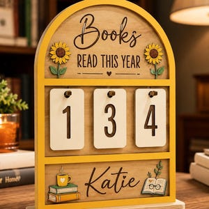 May include: An arched wooden book tracker sign with a yellow frame. The text reads Books READ THIS YEAR and Katie. It features two sunflowers, a stack of books with a mug, and an open book. Three white hanging tags display the number 134.