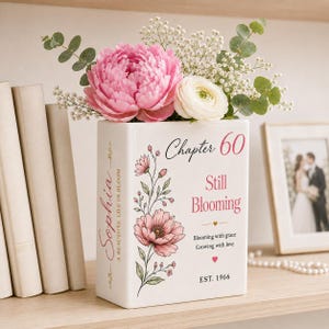 May include: A white ceramic book-shaped vase featuring pink floral illustrations and text that reads Chapter 60, Still Blooming, Sophia, A Beautiful Life in Bloom, Blooming with grace, Growing with love, and EST. 1966, filled with pink peonies and greenery.