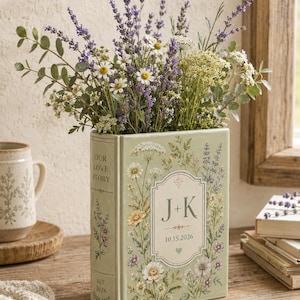 May include: A light green ceramic book-shaped vase featuring a floral print, the text "OUR LOVE STORY", "EST. 2026", and a central label with "J+K" and "10.15.2026". The vase is filled with a bouquet of purple lavender, white daisies, and green foliage.