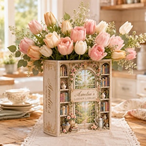 Puede incluir: Un florero rectangular con forma de libro titulado Amelia's Garden Library, que presenta un diseño de estantería en miniatura y ventana arqueada. El florero está lleno de un gran ramo de tulipanes rosas, melocotón y blancos.