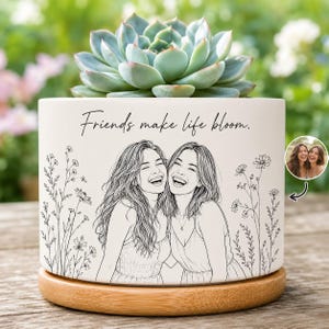 May include: A white ceramic cylindrical planter with a wooden base, containing a green succulent. The planter features a black line art illustration of two smiling people surrounded by wildflowers and the text, Friends make life bloom.