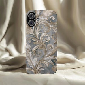 May include: A phone case featuring a detailed, ornate floral design in shades of gray and gold. The case has a glossy finish and a camera cutout. The design includes swirling leaves and floral patterns, creating an elegant aesthetic.