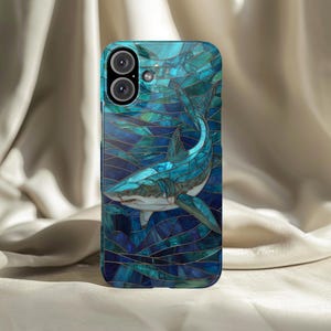 Può includere: Custodia per smartphone con un design a mosaico raffigurante uno squalo in tonalità blu e turchese. La custodia ha una finitura lucida e un'apertura per la fotocamera nella parte superiore.