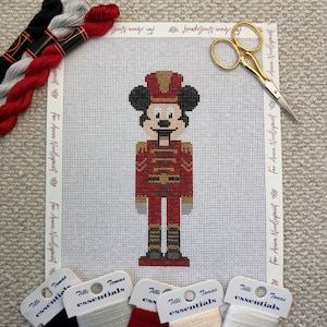 May include: Needlepoint canvas featuring a red and gold nutcracker design with Mickey Mouse ears. The canvas is surrounded by embroidery floss in black, red, and gray, along with gold scissors and thread labels.
