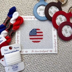 May include: A needlepoint canvas featuring a circular American flag design, surrounded by red, white, and blue embroidery floss. Several leather keychains in various colors are also present, along with a spool of white thread. The text "For Anna Needlepoint" is visible.