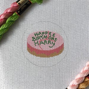 May include: A cross-stitch project featuring a pink cake design with the words "Happy Birthday Harry" in green. Pink and green embroidery floss are visible in the upper left and lower right corners, suggesting a crafting theme.