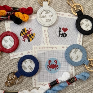 Puede incluir: Una colección de lienzos de punto de aguja con diseños temáticos de Maryland, incluyendo una bandera, un corazón con el texto MD y un cangrejo. El conjunto incluye varios llaveros circulares de cuero en blanco, negro, rojo y azul, junto con hilo de bordar.