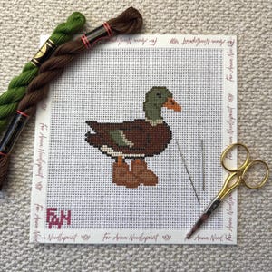 Może przedstawiać: Wyszywana krzyżykami kaczka z brązowymi, zielonymi i pomarańczowymi detalami na białym płótnie. Na płótnie widnieje napis "For Anna Needlepoint". Na zdjęciu znajdują się również dwie szpulki nici, igła i złote nożyczki.