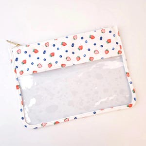 May include: A rectangular zippered pouch featuring a white fabric top section printed with a repeating pattern of small red strawberries and dark blue blueberries. The bottom section of the pouch is made of clear, transparent plastic material.