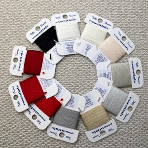 Könnte beinhalten: Eine Anordnung von farbigen Garnspulen in einem Kreis. Die Spulen sind mit der Marke "Tilli Tomas essentials" versehen und weisen verschiedene Farben auf, darunter Rot, Schwarz, Weiß und Beige. Das Garn ist auf kleinen Pappkarten aufgewickelt.