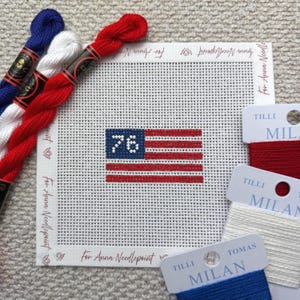 May include: Needlepoint canvas featuring a stitched American flag with the number "76" in the corner. Red, white, and blue embroidery floss and thread spools are arranged around the canvas. The text "For Anna Needlepoint" is printed on the canvas.