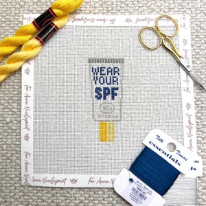 May include: A needlepoint canvas with a stitched design of a sunscreen tube that reads "WEAR YOUR SPF" in navy blue. The canvas is surrounded by yellow thread, gold scissors, and a spool of blue thread.