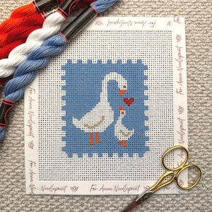 May include: Needlepoint canvas featuring two white geese with orange beaks and feet, and a red heart on a blue background. The canvas is surrounded by white fabric with text and is accompanied by red, white, and blue embroidery floss and gold scissors.