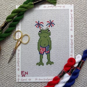 May include: Needlepoint canvas featuring a green frog wearing red, white, and blue swim trunks and fireworks on its head. The canvas is surrounded by green, red, white, and blue embroidery floss and gold scissors.