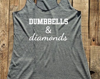 May include: Gray racerback tank top with the words "Dumbbells & diamonds" printed in white. The tank top is made of a soft material and has a scoop neck. The tank top is displayed on a rustic wooden surface.