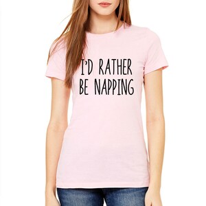 May include: A light pink t-shirt with the text "I'D RATHER BE NAPPING" in black, centered on the front. The shirt has short sleeves and a crew neck. The model is wearing blue jeans.