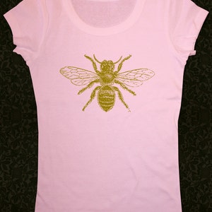 Op de afbeelding: Een roze T-shirt met een gouden bij-print op de voorkant.