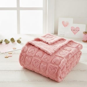 Könnte beinhalten: Eine gefaltete rosa Decke mit einem Herzmuster. Die Decke besteht aus einem weichen, plüschigen Material. Die Decke liegt auf einer weißen Holzoberfläche. Es gibt zwei Grußkarten mit Herzmotiven und dem Text "A Gift For You".