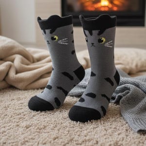 May include: A pair of gray cat-themed socks with black accents. The socks feature a cat face design on the front, with yellow eyes and white whiskers. The socks are on a beige carpet, with a fireplace in the background.